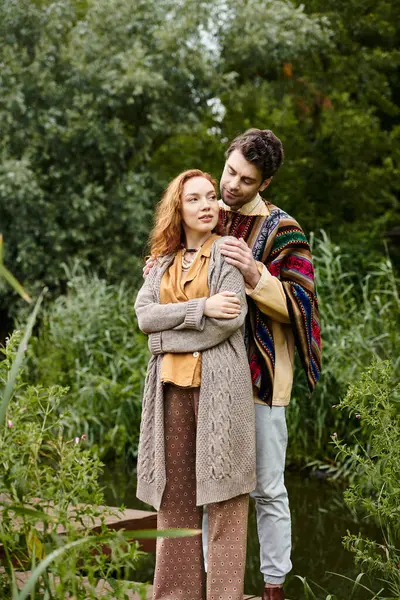 A man and woman in boho attire stand close together on a wooden bridge in a lush green park, sharing a tender moment. — Stock Photo