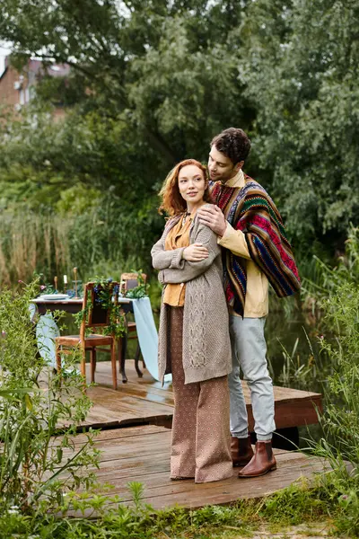 Un hombre y una mujer, vestidos con ropa de estilo boho, están en un muelle junto a un lago sereno. - foto de stock