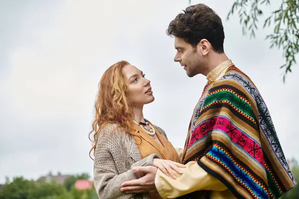 Un hombre y una mujer, vestidos al estilo boho, parados uno al lado del otro en un exuberante parque verde, en una cita romántica. - foto de stock