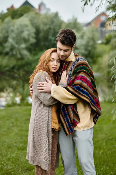 Un hombre y una mujer en traje boho se abrazan tiernamente en medio de la belleza natural de un parque verde, perdido en un momento de amor. - foto de stock