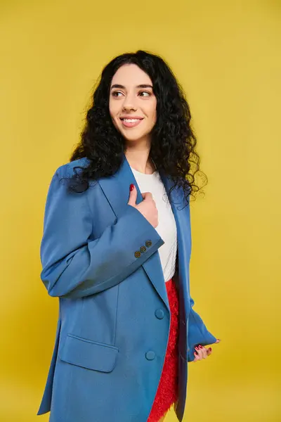 A young brunette woman with curly hair strikes a pose in a stylish blue jacket, exuding confidence and elegance against a yellow background. — Stock Photo