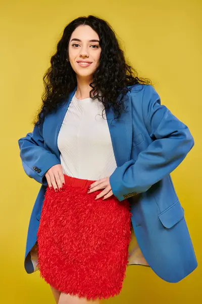 A young brunette woman with curly hair posing in a stylish blue jacket and red skirt against a yellow studio backdrop. — Stock Photo