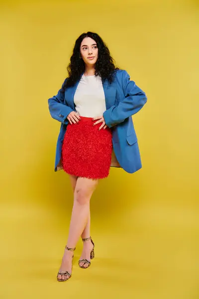 Young brunette woman with curly hair poses confidently in a blue jacket and red skirt against a vibrant yellow backdrop. — Foto stock