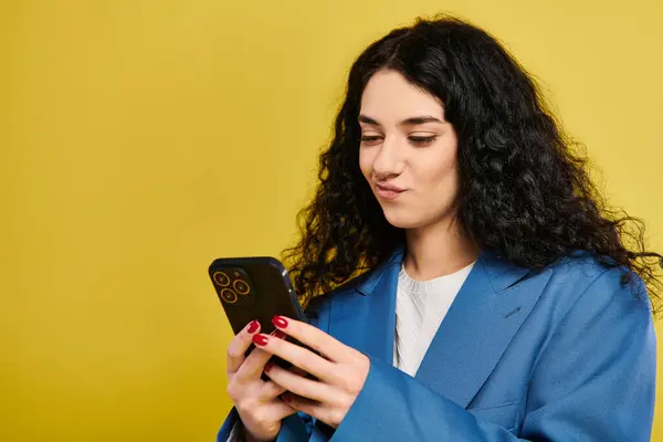 Una giovane donna elegante con i capelli ricci in una giacca blu assorbita nel suo telefono cellulare su uno sfondo giallo. — Foto stock
