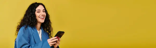 Una giovane donna bruna espressiva con i capelli ricci che sorride mentre tiene in mano uno smartphone, trasudando gioia in un ambiente da studio con uno sfondo giallo. — Foto stock