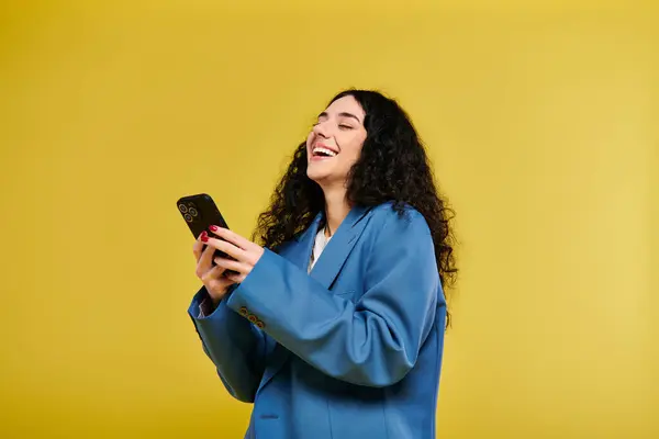 Una mujer joven y elegante con el pelo rizado en una chaqueta azul sostiene un teléfono celular sobre un fondo amarillo brillante. - foto de stock