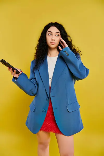 A young brunette woman with curly hair, dressed in a blue jacket, holding a cell phone in a vibrant yellow studio. — Stock Photo