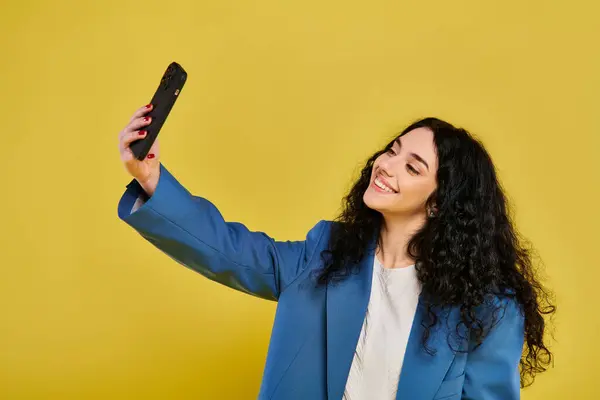 Une jeune femme brune aux cheveux bouclés, vêtue élégamment, sourit en prenant un selfie sur son téléphone portable dans un studio au fond jaune. — Photo de stock