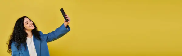 A brunette woman in stylish attire poses with curly hair, capturing her emotions with a cell phone against a yellow backdrop. — Stock Photo