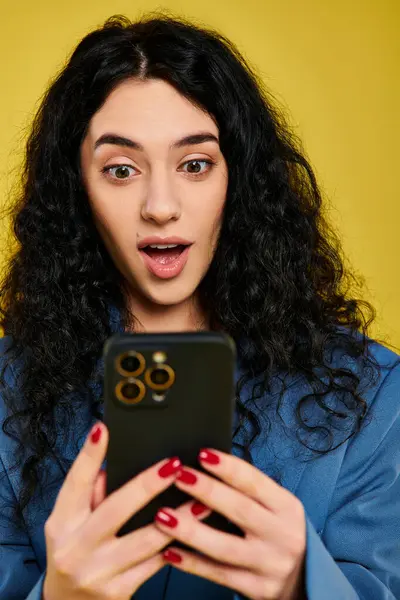 A young brunette woman with curly hair, dressed stylishly, holds a cell phone with a surprised expression against a yellow backdrop. — Stock Photo