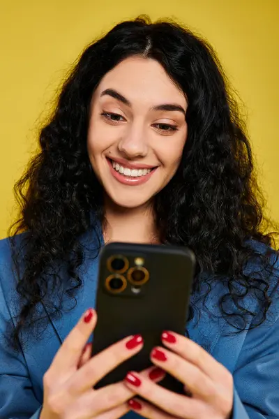 Une jeune femme brune aux cheveux bouclés en tenue élégante exprimant des émotions tout en tenant un téléphone portable, sur fond jaune. — Photo de stock