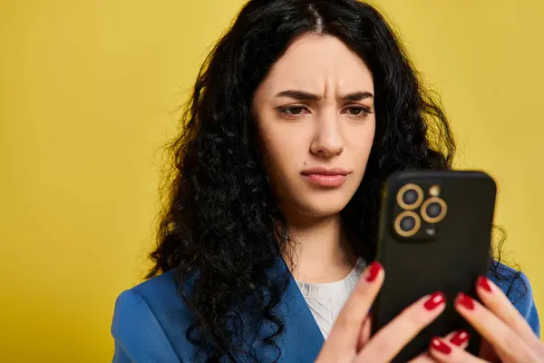 Eine junge brünette Frau mit lockigem Haar in stylischer Kleidung hält ein Handy in der Hand und zeigt verschiedene Emotionen vor gelbem Hintergrund. — Stockfoto