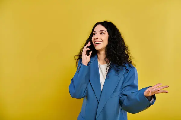 Jeune femme brune aux cheveux bouclés en veste bleue parlant sur téléphone portable dans une pose élégante sur fond jaune. — Photo de stock