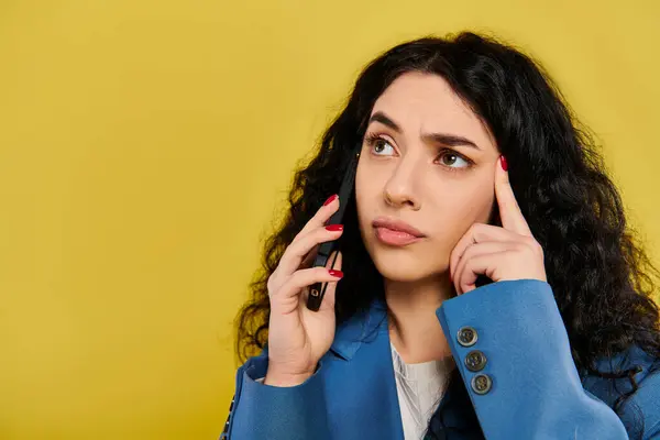 Una mujer morena con el pelo rizado con un atuendo elegante sostiene un teléfono celular en su oído, mostrando varias emociones sobre un fondo amarillo. - foto de stock