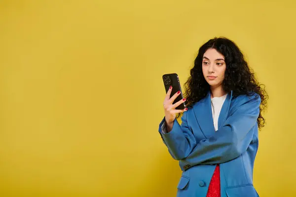 Jeune femme brune aux cheveux bouclés, vêtue d'une veste bleue, regardant son téléphone portable dans un studio au fond jaune. — Photo de stock