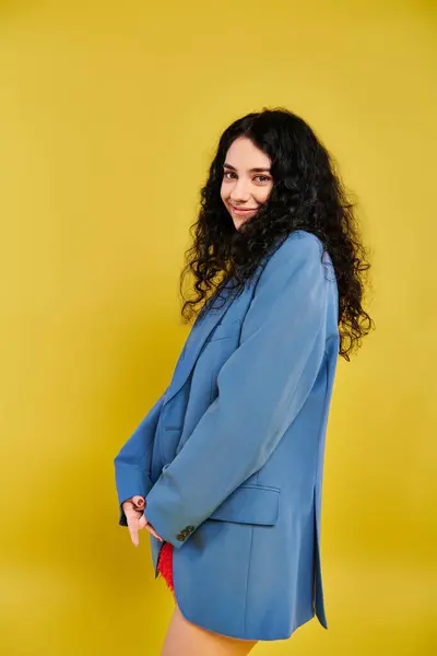 Eine junge brünette Frau mit lockigem Haar, in einem blauen Anzug, posiert elegant in einem Studio mit gelbem Hintergrund. — Stockfoto