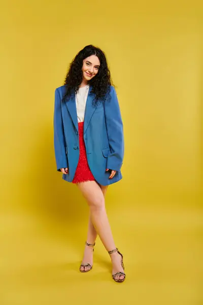 Una mujer joven y elegante con el pelo rizado posa en una chaqueta azul y falda roja, mostrando su sentido único de la moda en un fondo amarillo. - foto de stock