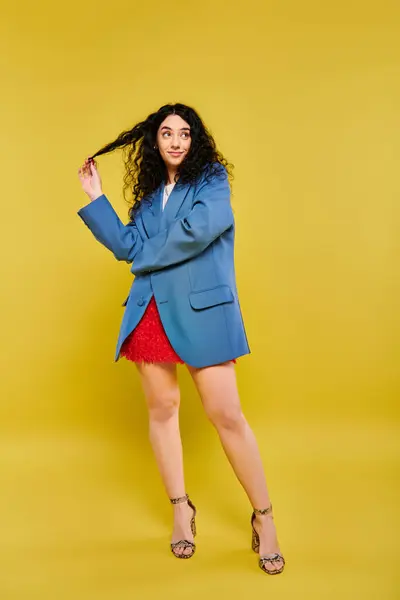 A brunette woman with curly hair posing in a blue jacket and red skirt, exuding style and charm against a vibrant yellow backdrop. — Stock Photo