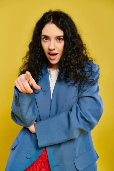 A young, brunette woman with curly hair striking a pose in a blue jacket while pointing directly at the camera. — Stock Photo