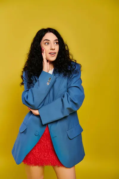 A young, brunette woman with curly hair poses for a picture in a stylish blue jacket, showcasing her emotions against a yellow background. — Stock Photo