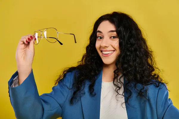 A young, curly-haired brunette woman showcasing a pair of glasses while exuding style and sophistication against a vibrant yellow backdrop. — Stock Photo