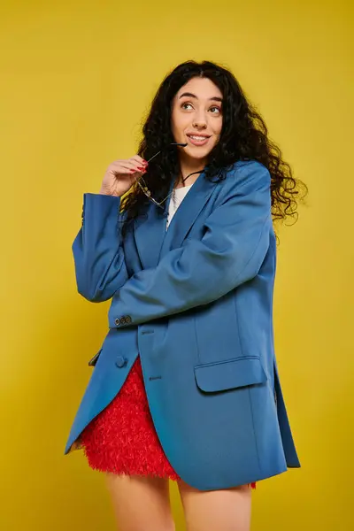 A young brunette woman with curly hair poses in a stylish blue jacket and red skirt, expressing emotions against a yellow background. — Stock Photo