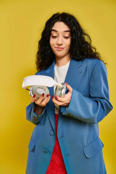 A young brunette woman with curly hair poses in a studio setting, showcasing her emotions as she holds a white headphones in her hands against a yellow background. — Stock Photo