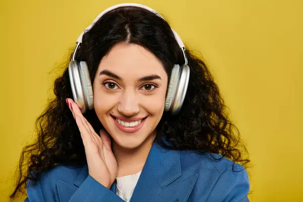 A young brunette woman, with curly hair and stylish attire, smiles while wearing headphones in a studio with a yellow background. — Stock Photo