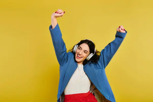 Brunette woman with curly hair poses confidently, raising her hands in a blue jacket against a vibrant yellow studio backdrop. — Stock Photo