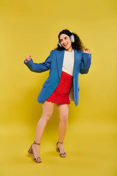 Femme brune aux cheveux bouclés pose dans une veste bleue élégante et une jupe rouge, exsudant confiance et élégance sur un fond jaune. — Photo de stock