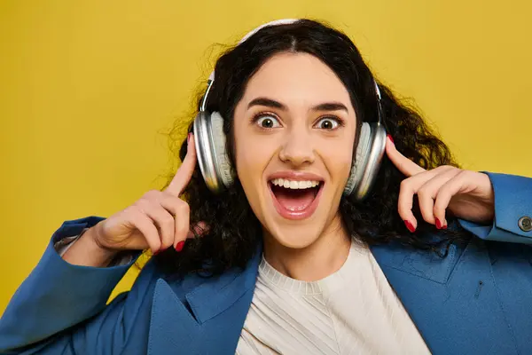 Una joven morena de pelo rizado con un atuendo elegante hace una cara divertida mientras usa auriculares en un estudio con un fondo amarillo. - foto de stock