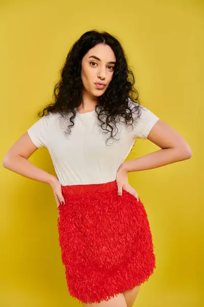 A young brunette woman with curly hair strikes a pose in a stylish red skirt against a yellow studio background. — Stock Photo