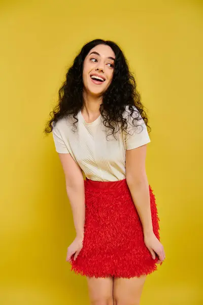 Young brunette woman with curly hair striking a pose in a vibrant red skirt against a yellow studio backdrop. — Stock Photo