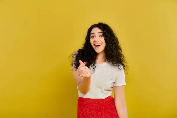 Giovane donna bruna con i capelli ricci in posa in una camicia bianca e gonna rossa, trasudante stile ed emozione su uno sfondo giallo vibrante. — Foto stock