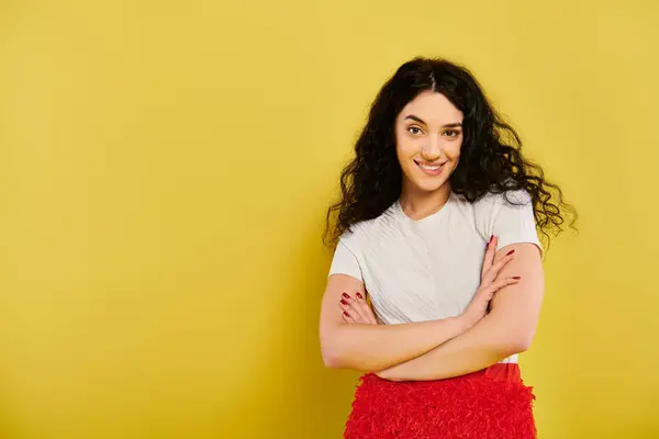 A young brunette woman with curly hair stands confidently with arms crossed against a vibrant yellow backdrop. — Stock Photo