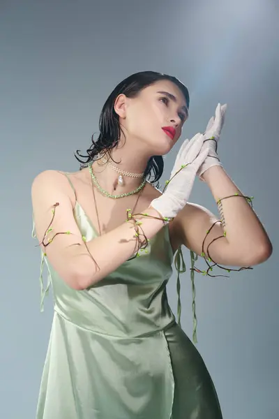 A young, beautiful woman with red lips poses elegantly in a green dress and white gloves in a studio setting on a grey background. — Stock Photo