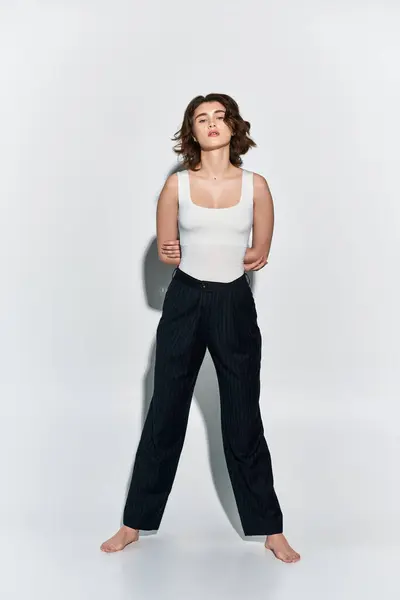 Eine hübsche junge Frau in schwarzer Hose und weißem Tank-Top steht selbstbewusst vor einer weißen Wand in einem Studio-Ambiente.. — Stockfoto