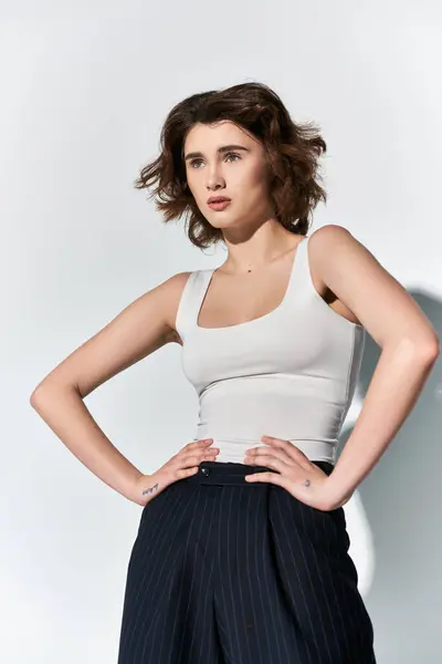 A stylish young woman strikes a strong pose, hands on hips, in black pants and white tank top, set against a grey backdrop. — Stock Photo