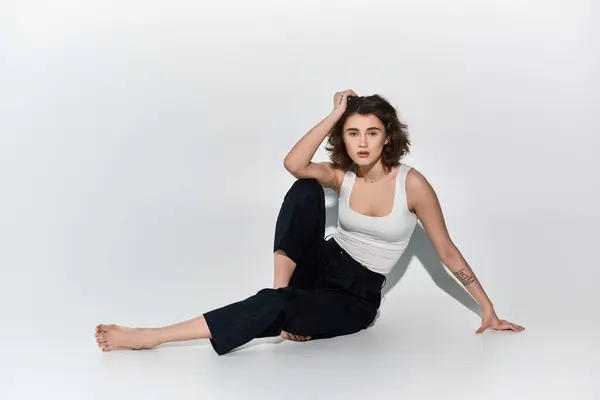 A pretty young woman sitting gracefully on the ground with her legs crossed, dressed in black pants and a white tank top. — Stock Photo