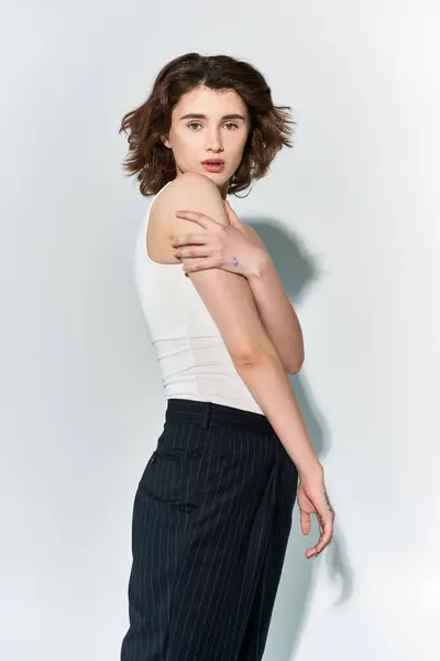A pretty young woman in a white tank top and black pants strikes a graceful pose in a studio setting with a grey background. — Stockfoto