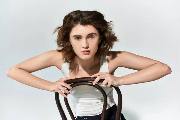 Eine hübsche junge Frau posiert selbstbewusst in einem Studio, trägt eine schwarze Hose und ein weißes Tank-Top und hält einen braunen Stuhl in der Hand — Stockfoto