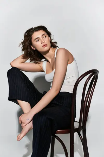 A pretty young woman in black pants and a white tank top sitting gracefully atop a wooden chair in a studio setting against a grey background. — Stock Photo