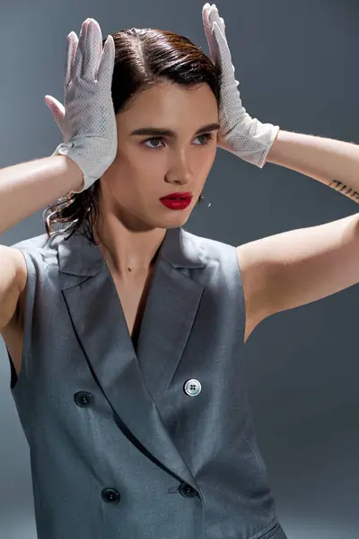 Stylish young woman poses in an elegant gray suit with a vest and white gloves in a studio setting on a grey background. — Stock Photo
