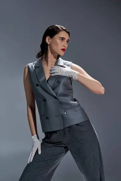 A stylish young woman poses in an elegant gray suit with white gloves in a studio setting against a gray background. — Stock Photo