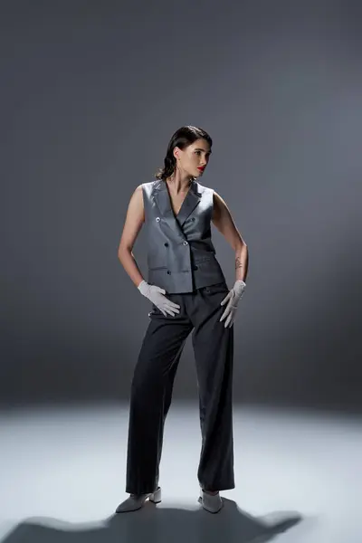 A stylish young woman elegantly poses in a gray suit with a vest and white gloves in a studio setting on a grey background. — Stock Photo