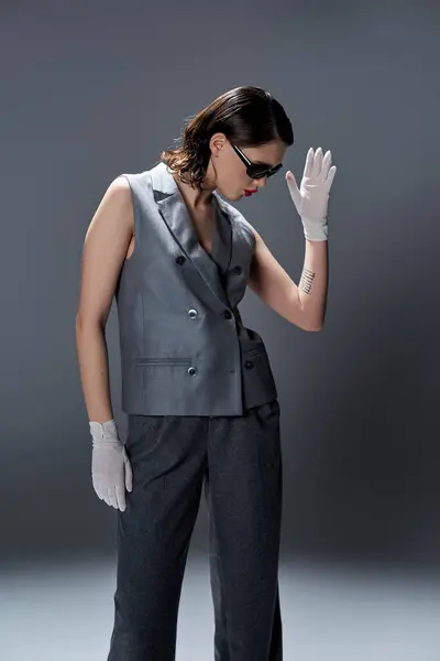 Eine stilvolle junge Frau posiert elegant in einem grauen Anzug mit Weste und weißen Handschuhen in einem Studio vor grauem Hintergrund.. — Stockfoto