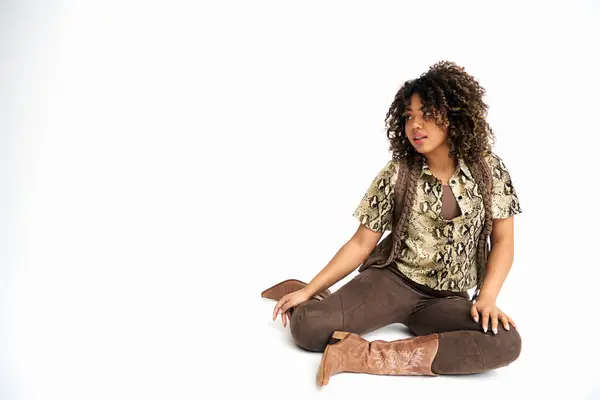Stylische afrikanisch-amerikanische Frau in schicker Kleidung sitzt auf dem Boden vor weißem Hintergrund und schaut weg — Stockfoto