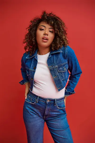 Appealing chic african american woman in denim attire posing on red backdrop and looking at camera — Stock Photo