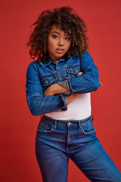 Alluring chic african american woman in denim attire posing on red backdrop and looking at camera — Stock Photo