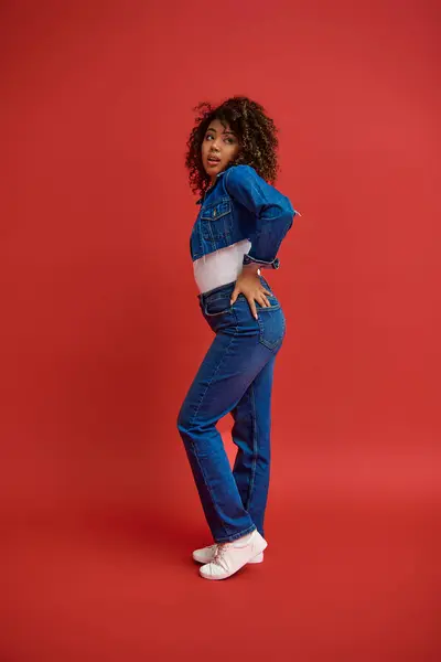 Витончена молода афроамериканка в стильному джинсовому вбранні, дивлячись на червоний фон — стокове фото